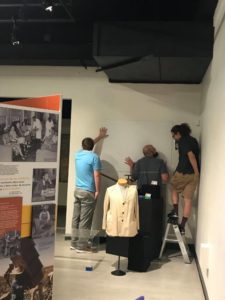 Three people standing by a wall to hang items. One is on a ladder